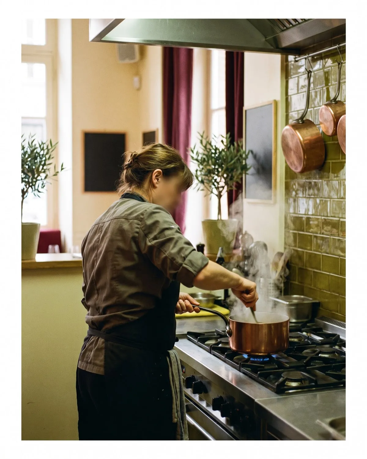Margot, la cheffe, de dos devant son fourneau, tablier noué, une casserole en cuivre à la main