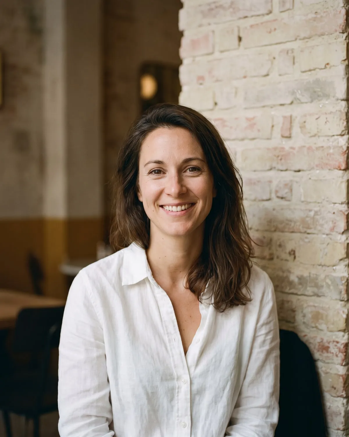 Une femme en chemise blanche, souriante, devant un mur de brique clair, cadrage buste