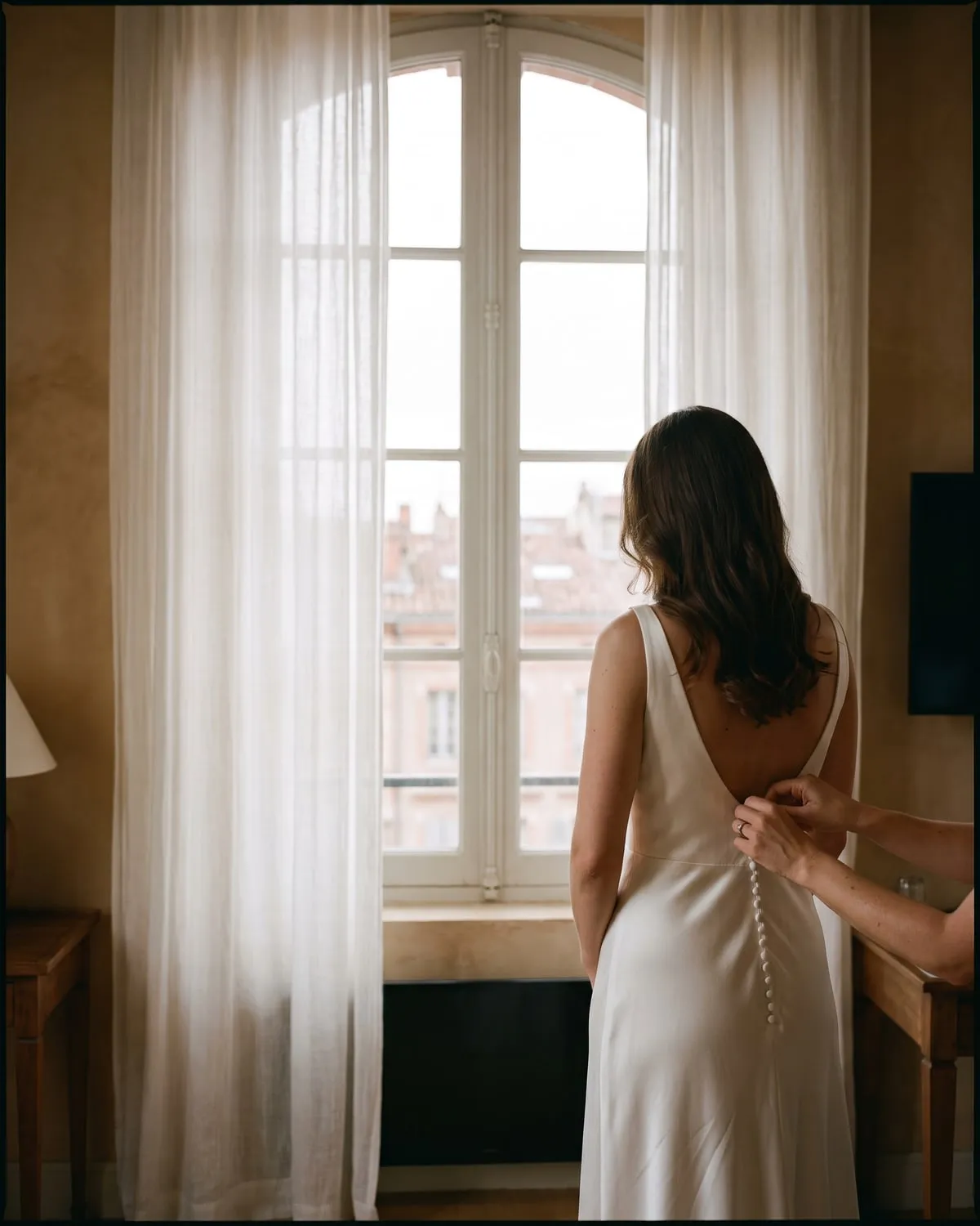 Une mariée de dos, devant une fenêtre, pendant qu’on ferme les boutons de sa robe