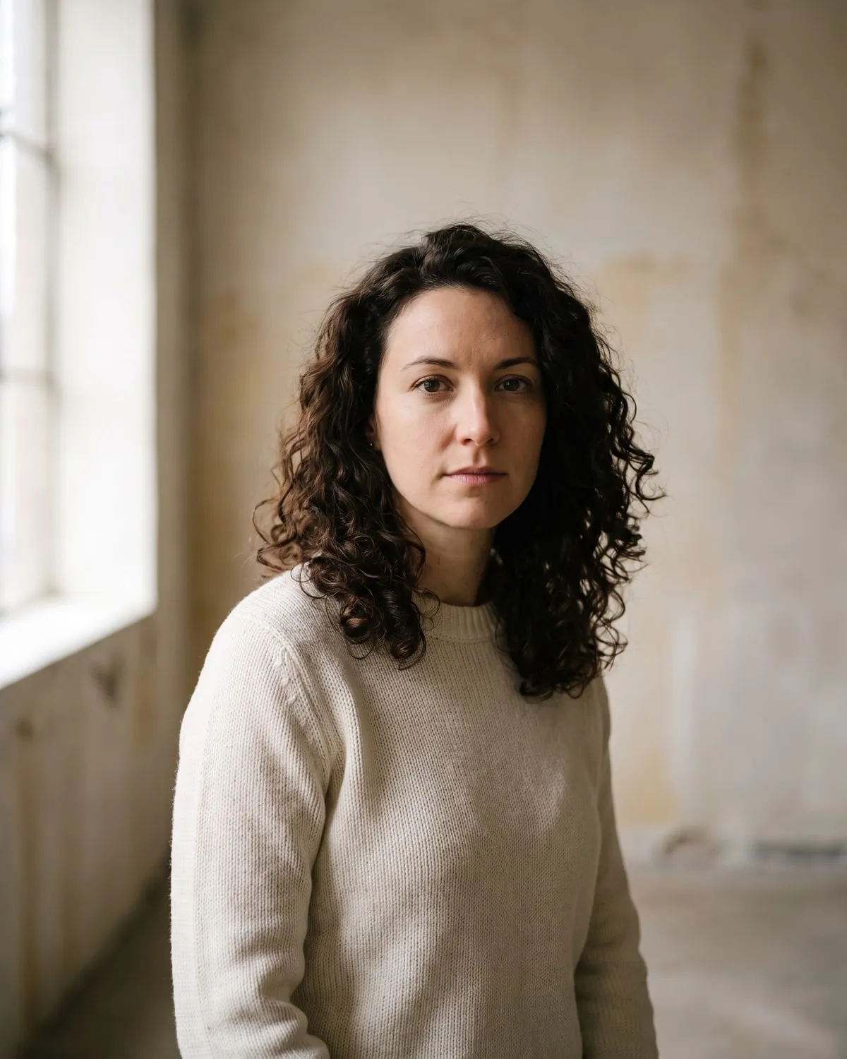 Portrait d’une femme aux cheveux bouclés, de trois quarts, devant un mur clair, lumière de fenêtre